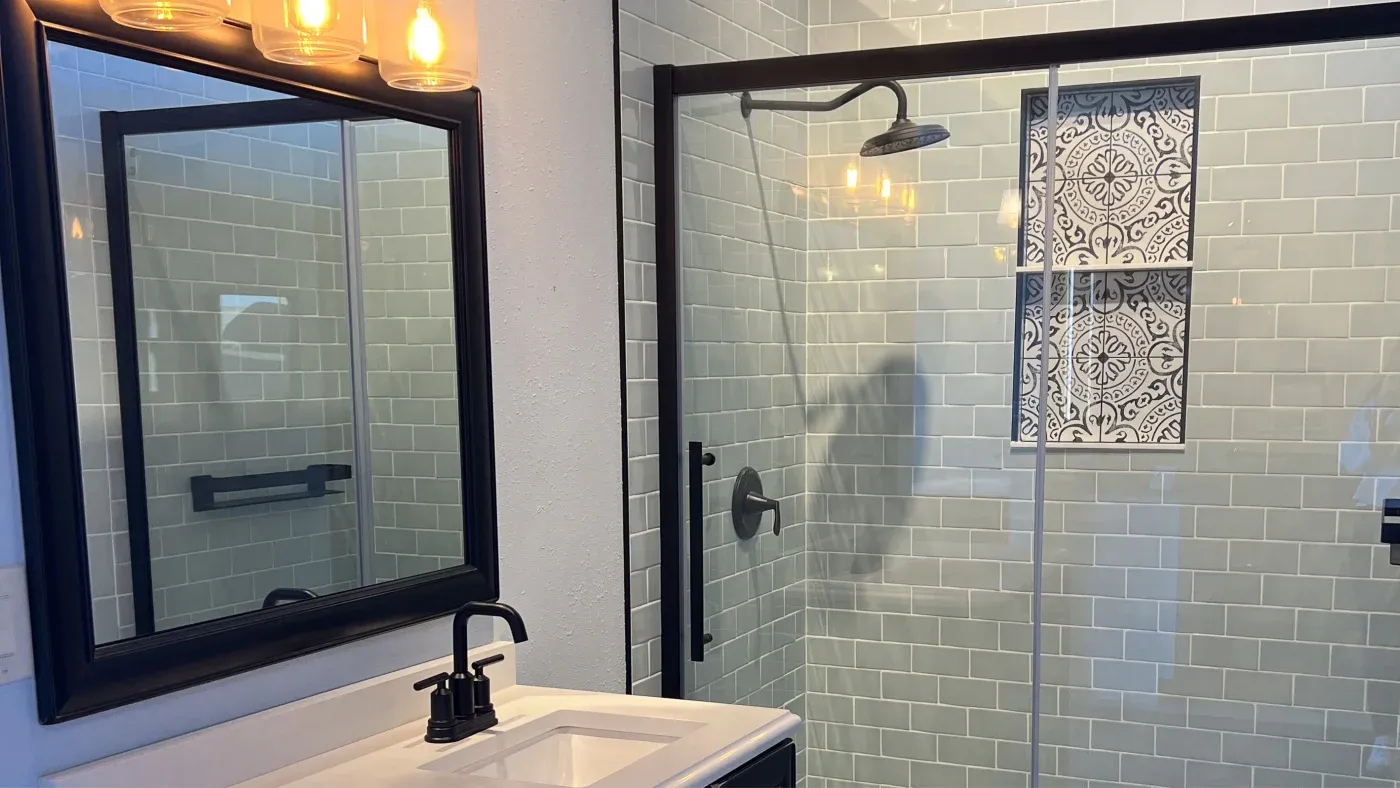 A bathroom with a walk-in shower that has glass doors and green tiles with an accent tile shelf